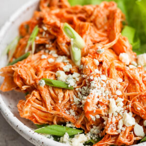 Shredded slow cooker buffalo chicken on a plate with blue cheese crumbles and sliced green onions.
