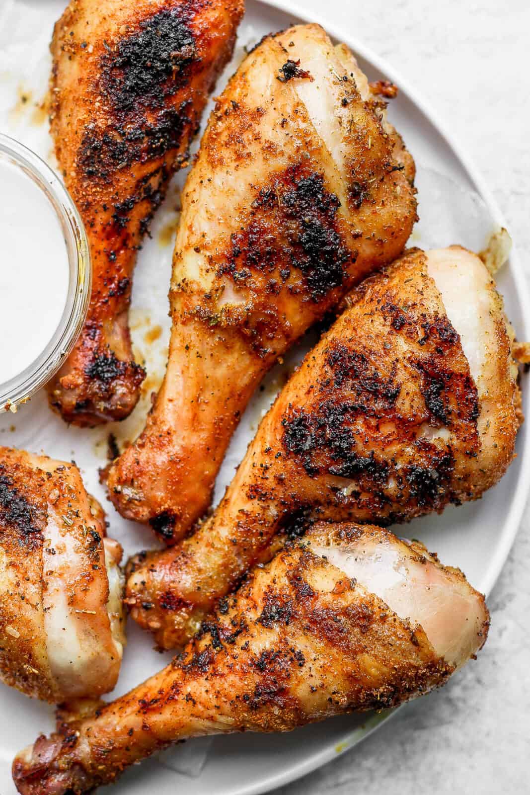 A plate with five grilled chicken legs and a little bowl of ranch dressing. 