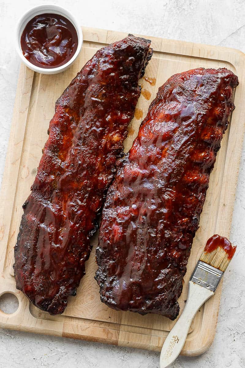 A wooden cutting board with two racks of ribs on top as well as a little bowl of bbq sauce and a small brush.