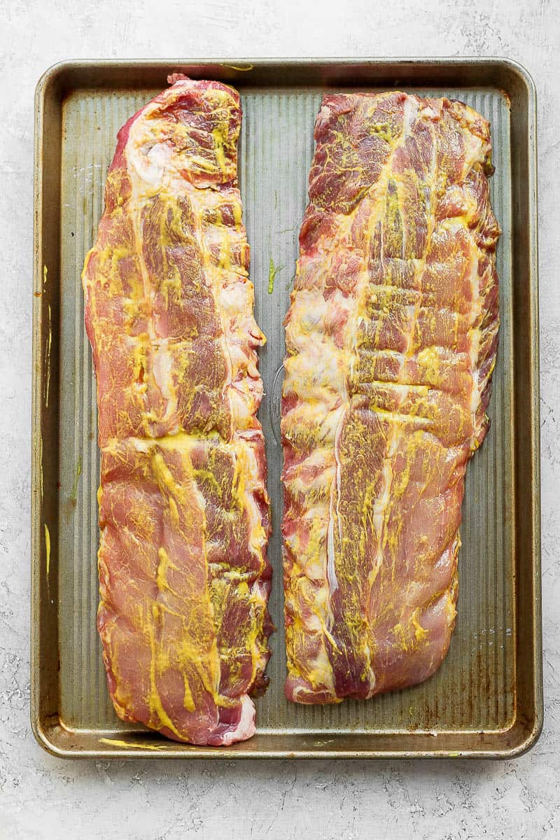 Two racks of ribs on a baking sheet covered in yellow mustard. 