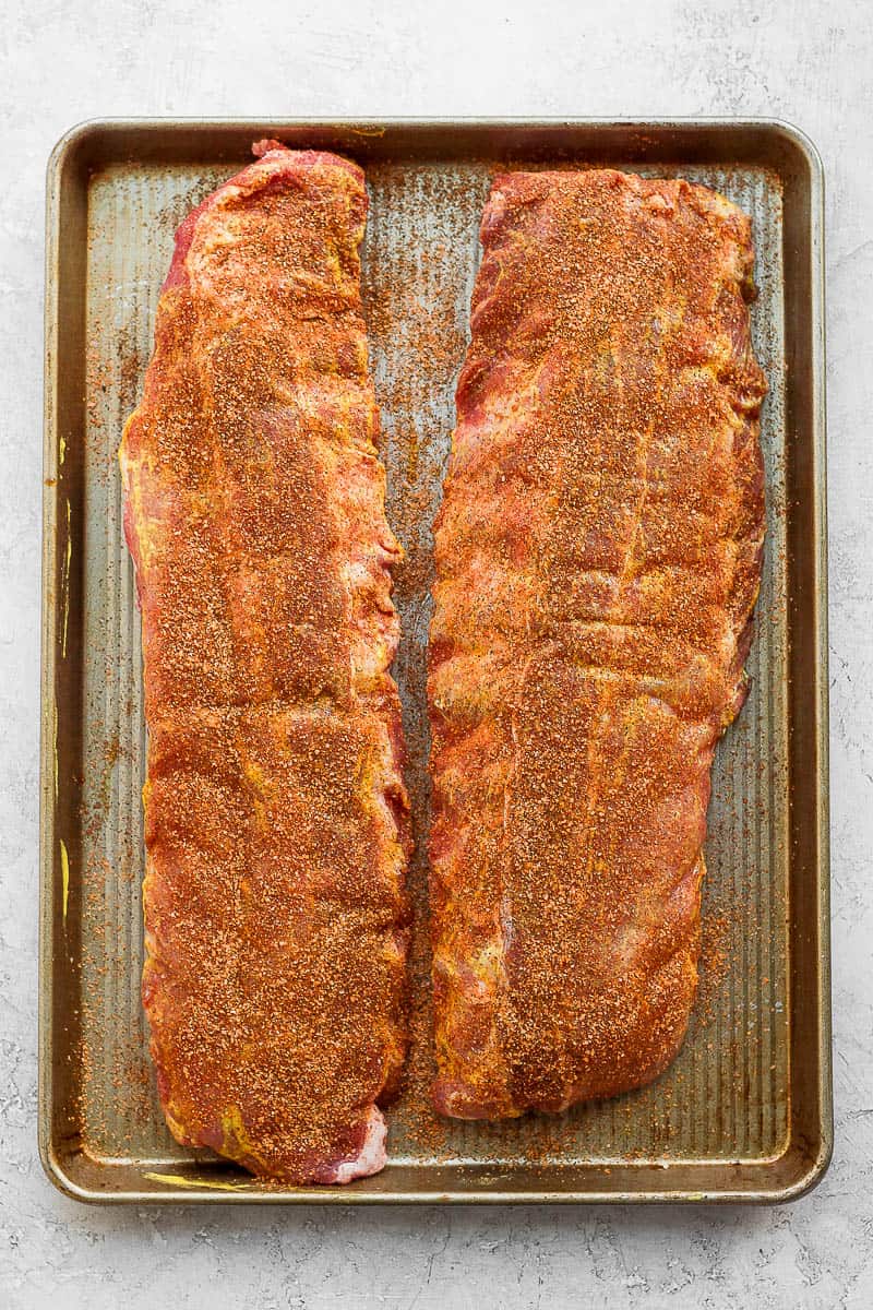 Two racks of ribs on a baking sheet covered in dry rub.