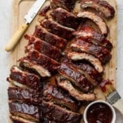 A wooden cutting board with 2 racked of oven baked ribs on top, sliced with a bowl of bbq sauce next to them.