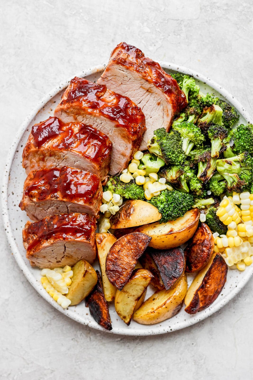 Pork tenderloin, sliced, on a plate with grilled broccoli, corn and potato wedges. 