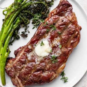 A white plate with a reverse seared steak topped with butter and roasted broccolini on the side.