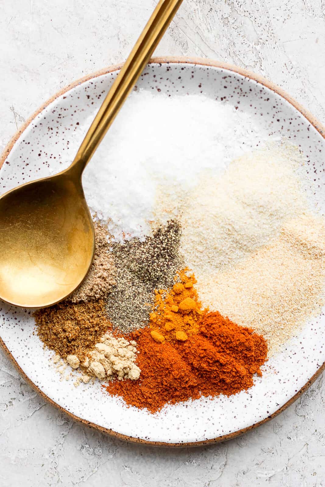 A speckled plate with all the different spices on it with a golden spoon.