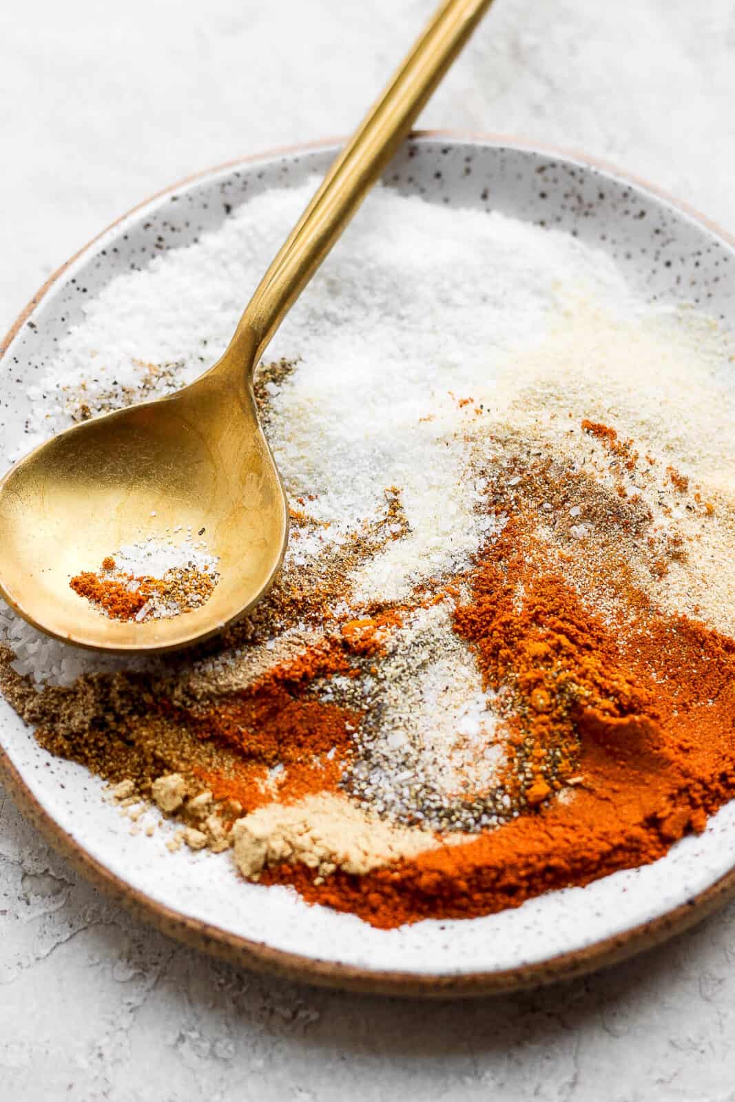 A speckled plate with all the different spices on it with a golden spoon.