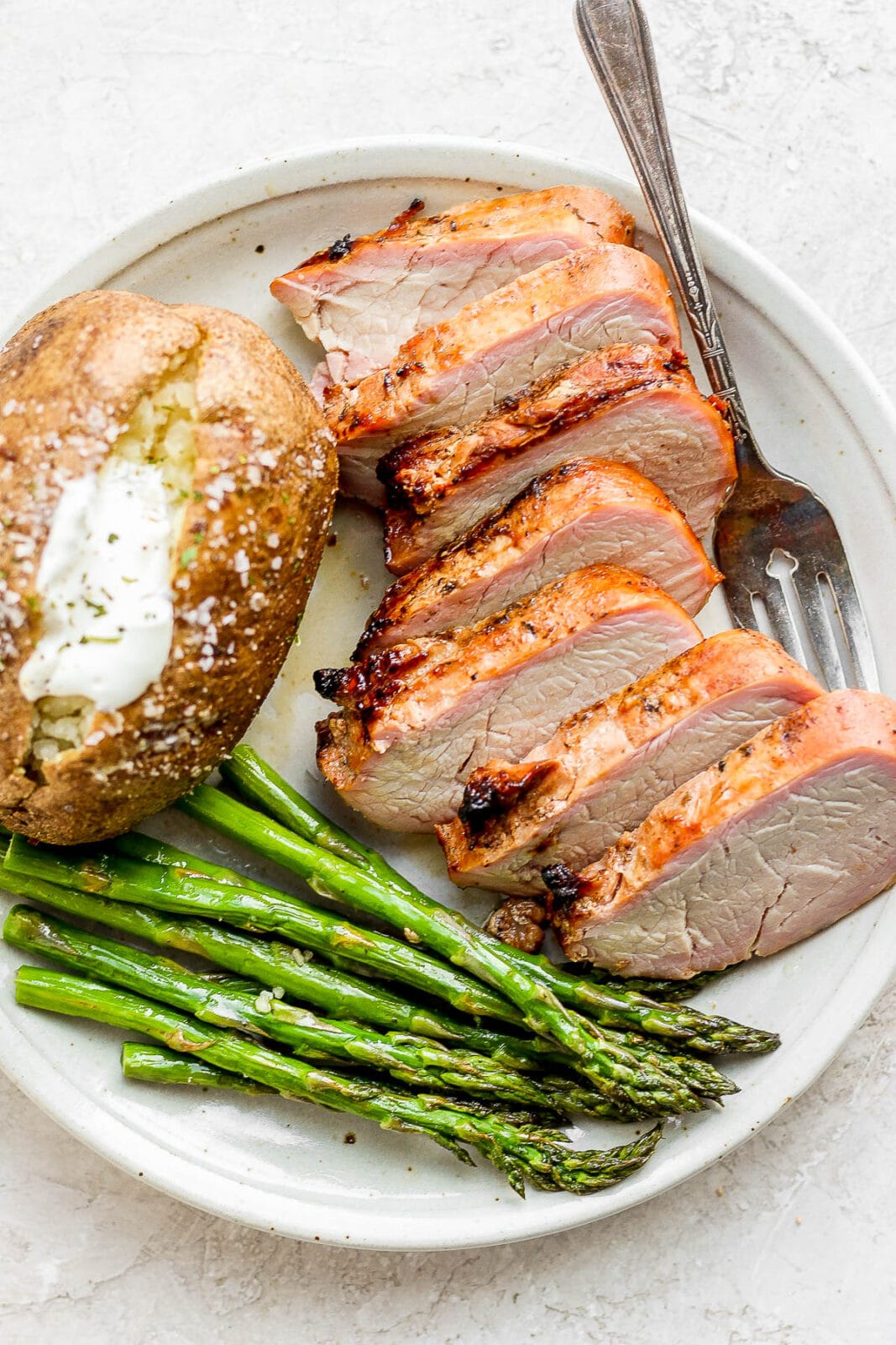 A smoked pork tenderloin on a plate with asparagus and a baked potato. 