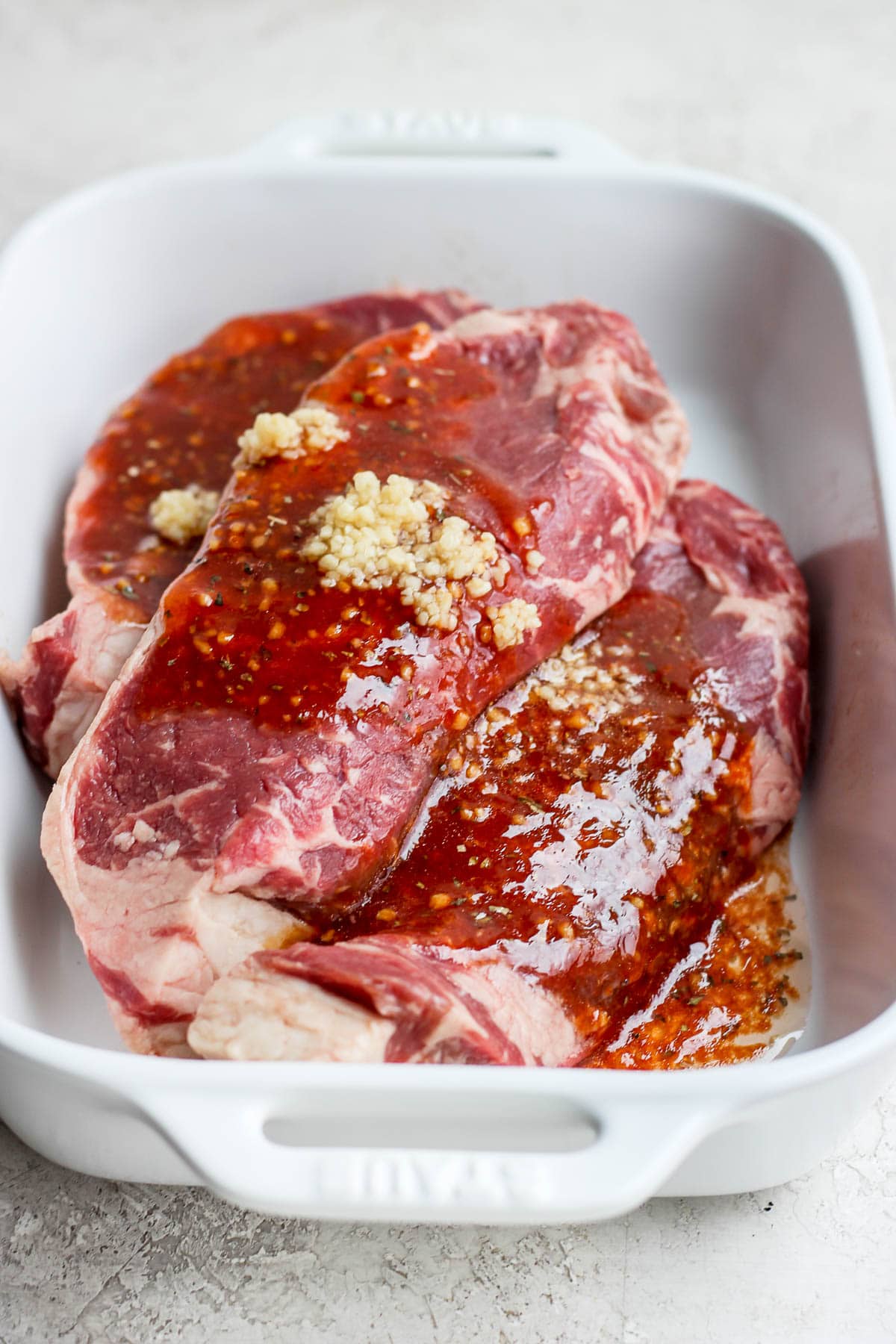 Three steaks in a white casserole dish with steak marinade on top of them. 