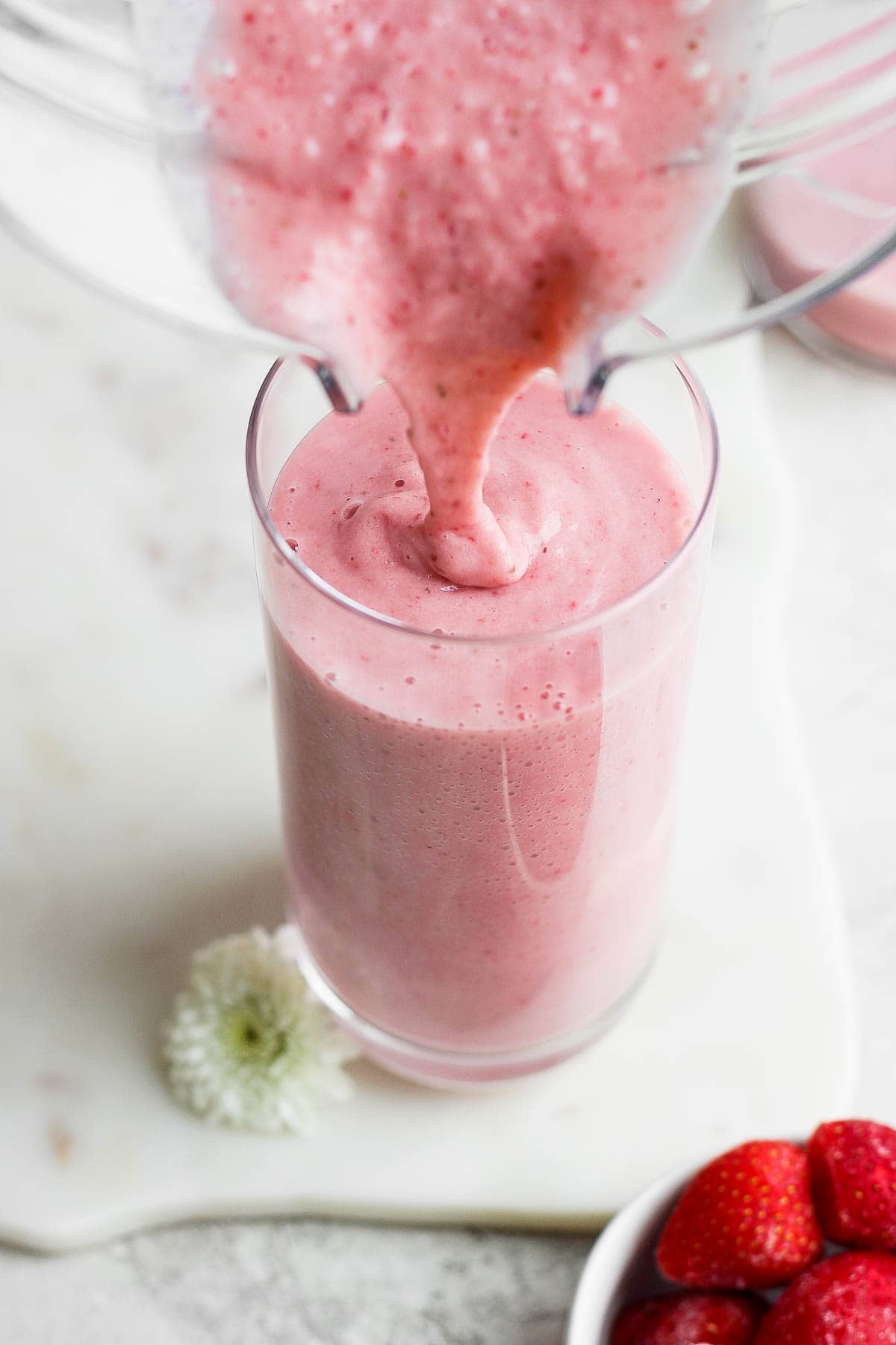 Strawberry smoothie being poured out of a blender into a tall glass.
