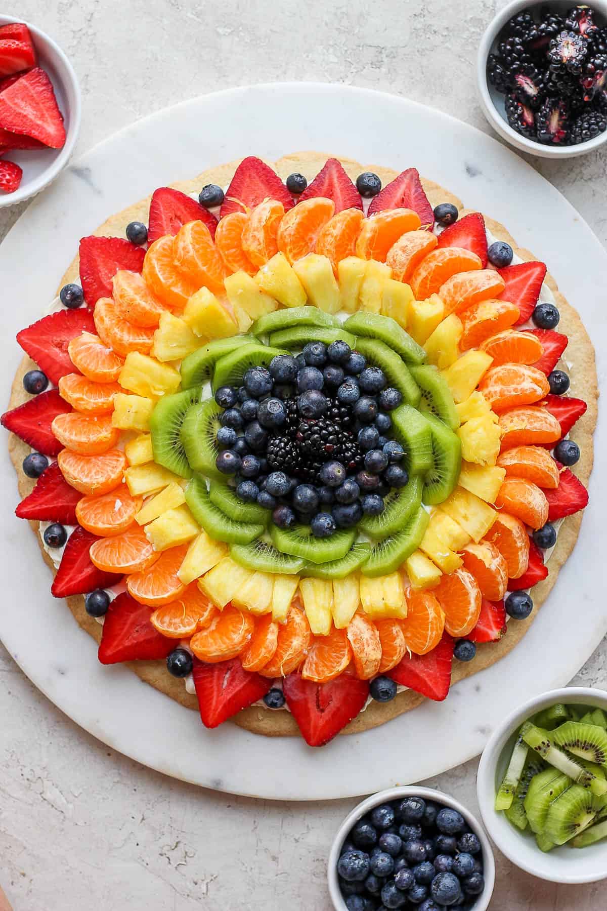 Fruit pizza on a white marble board with little bowls of fruit around it. 