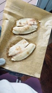 Someone holding a parchment lined baking sheet with a oven mitt on their hand and baked tilapia filets on the pan.