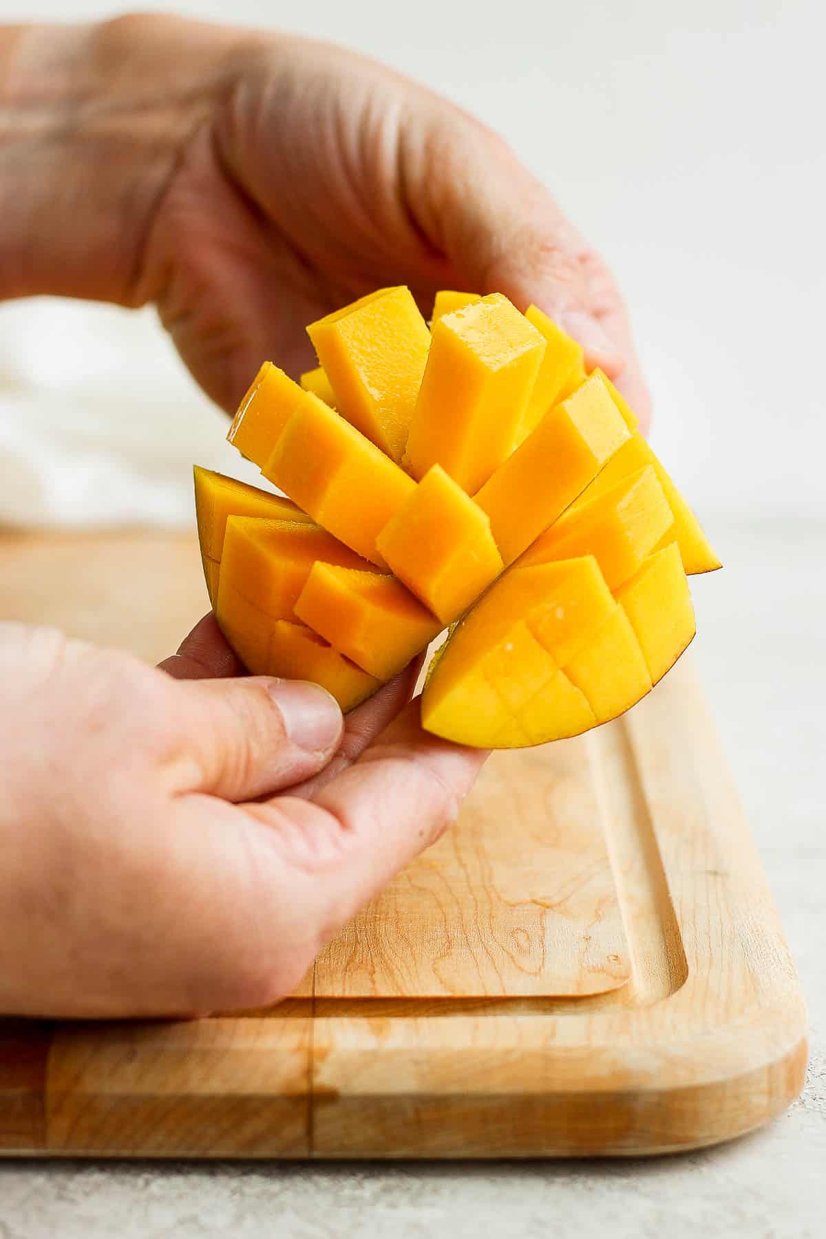 A hand pushing the rounded side of the mango to show the cubes attached to the skin.