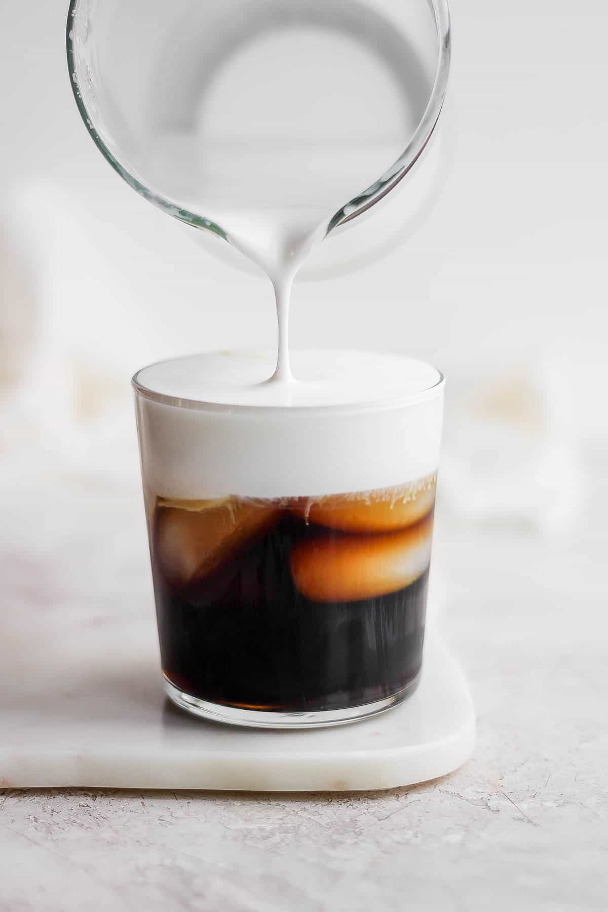 Someone pouring cold foam into a glass of cold brew coffee. 