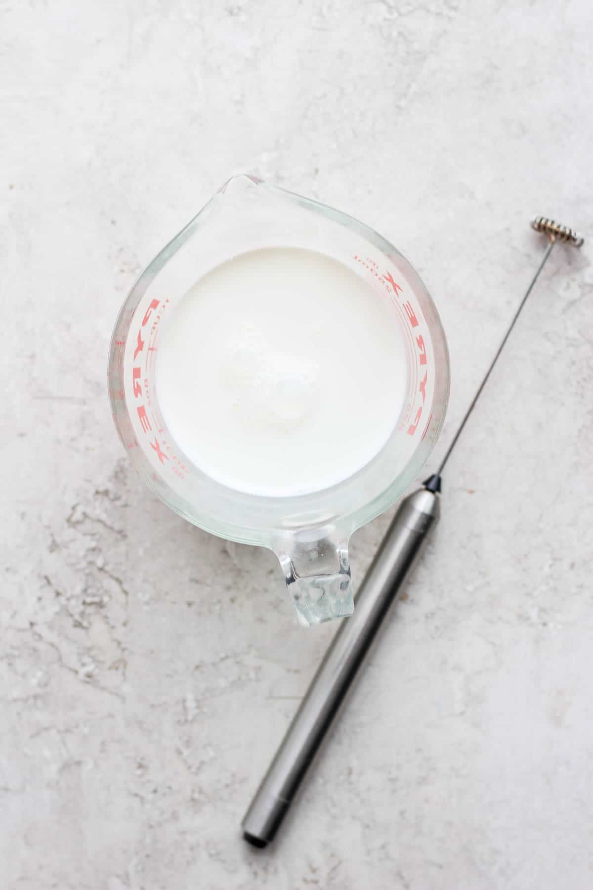 A handheld frother next to a glass measuring cup full of milk. 