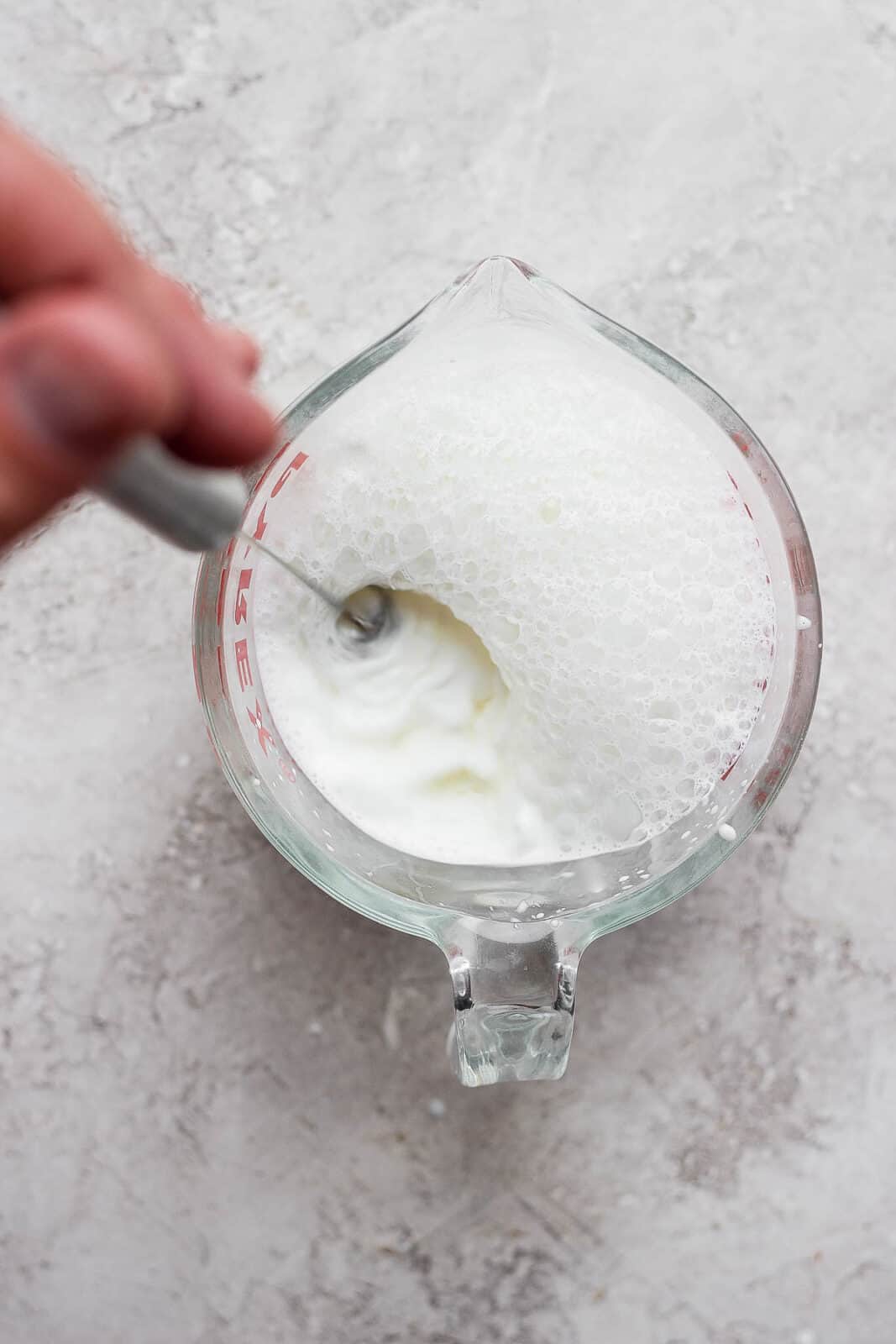 Top shot of someone using a handheld frother to froth milk in a glass measuring cup. 