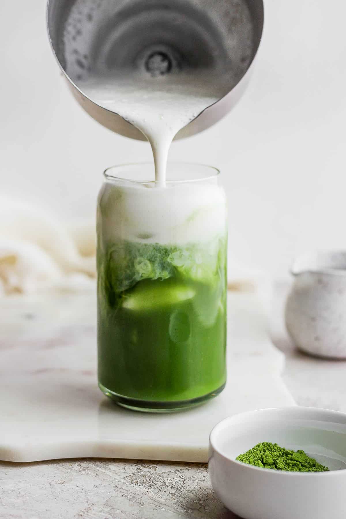 Someone pouring cold foam into a glass of iced matcha latte. 