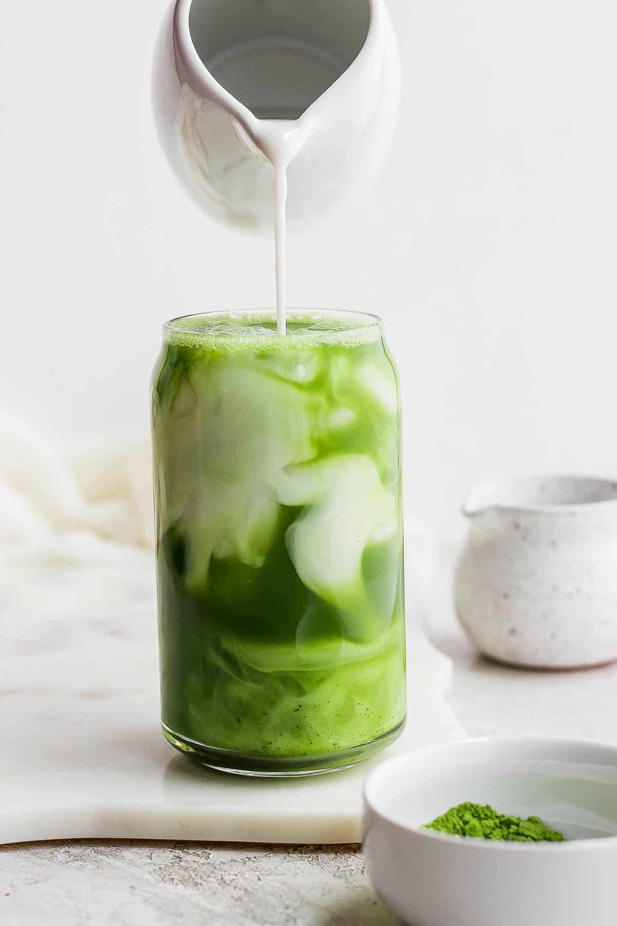 Someone pouring almond milk into a glass of iced matcha. 