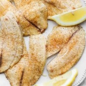 Baked tilapia filets on a white plate with some lemon wedges.