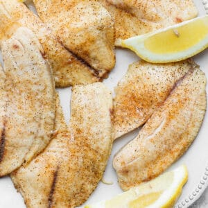 Baked tilapia filets on a white plate with some lemon wedges.