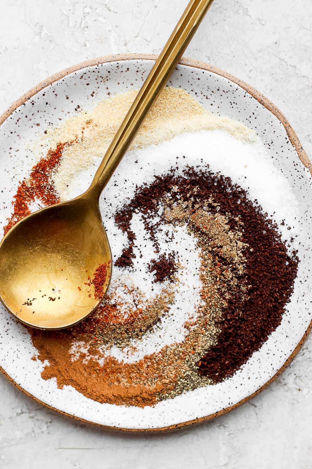 A plate with ingredients for steak dry rub swirled together with a golden spoon next to them. 