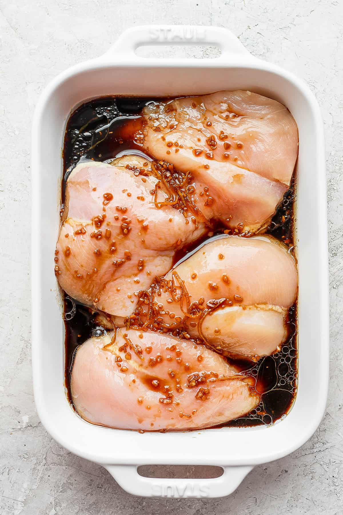 Raw chicken breasts in a pan with marinade on top.