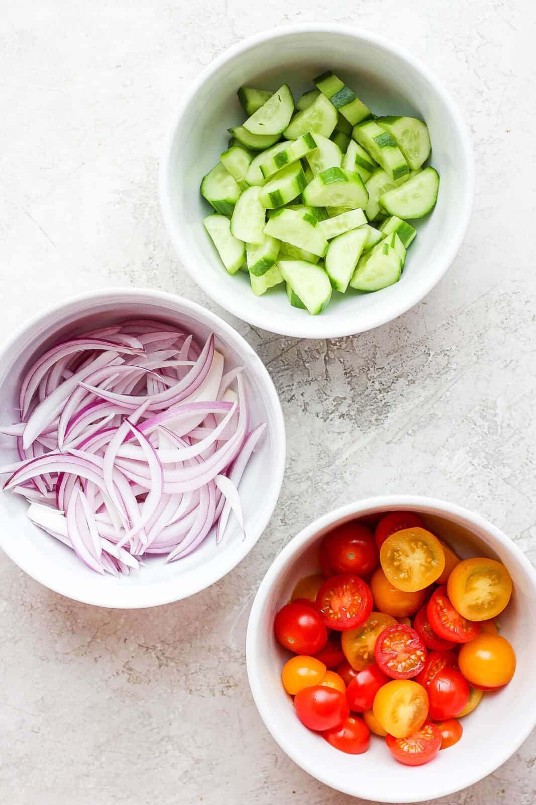 Three bowls of different ingredients for grilled salmon salad: cucumber, red onion and cherry tomatoes. 