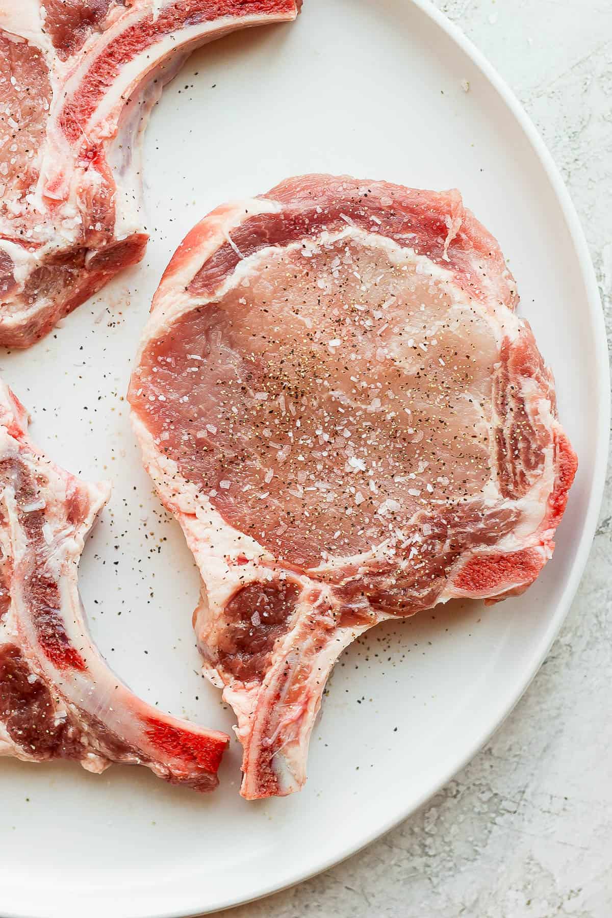 Raw pork chops on a plate with salt and pepper sprinkled on top.