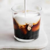 A glass of coffee with sweet cream cold foam being poured in it.