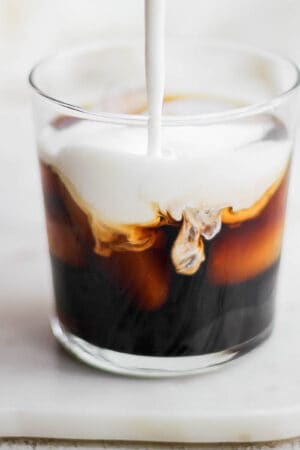 A glass of coffee with sweet cream cold foam being poured in it.