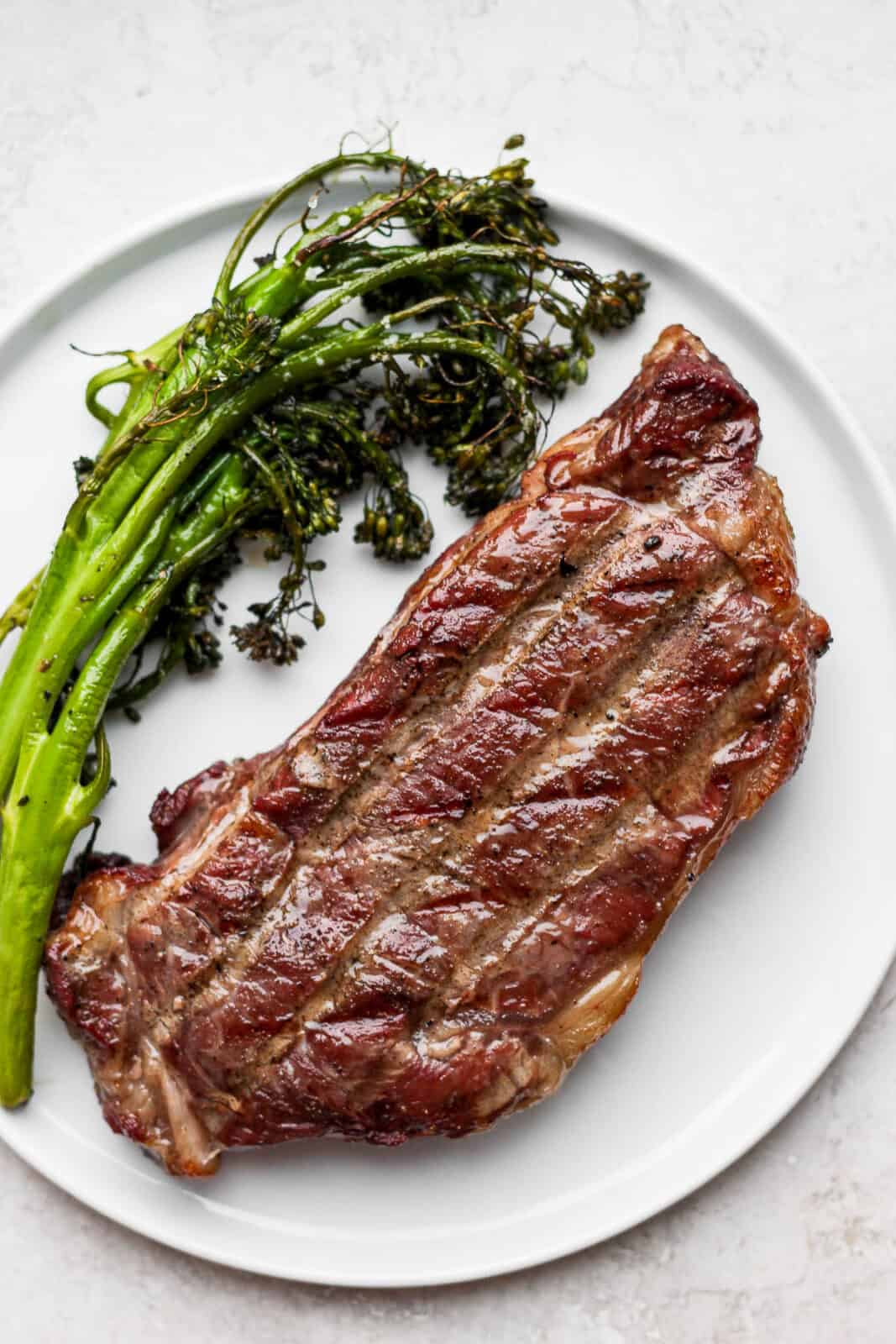 A plate with a reverse seared steak covered in melted butter and parsley with some grilled broccolini next to it.
