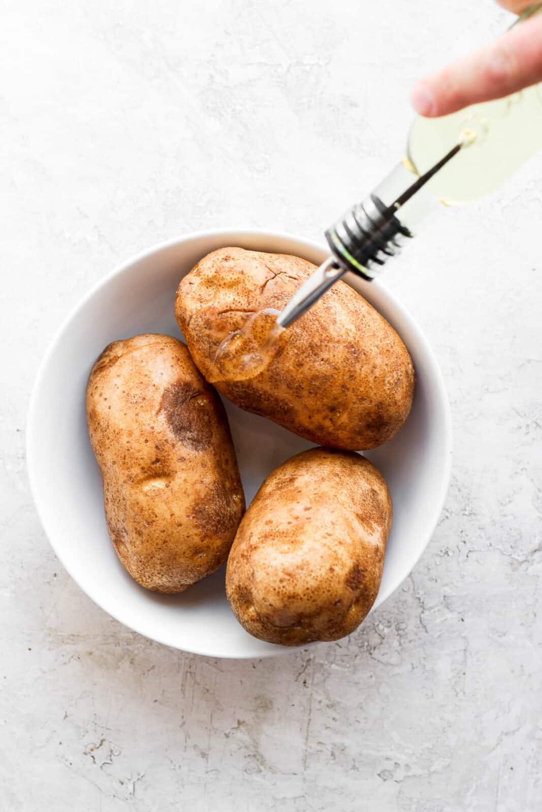Three potatoes in a white bowl being drizzled with oil.