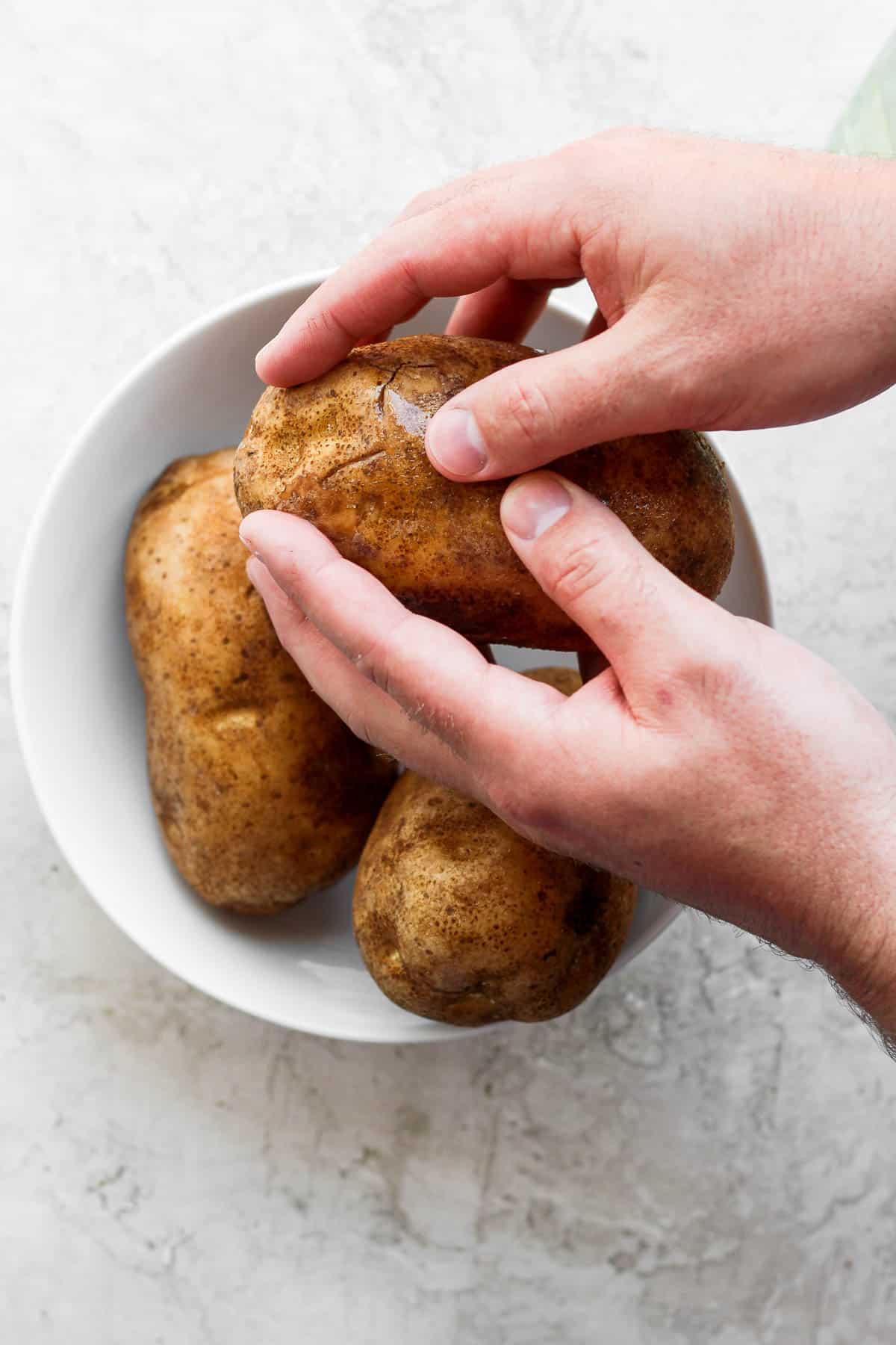 Two potatoes in a white bowl while one is being rubbed with olive oil by hands.