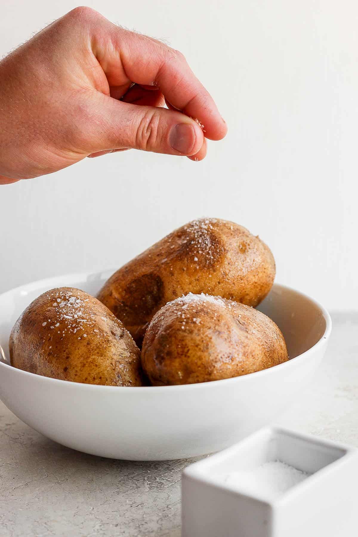 Three potatoes in a white bowl being sprinkled with kosher salt.