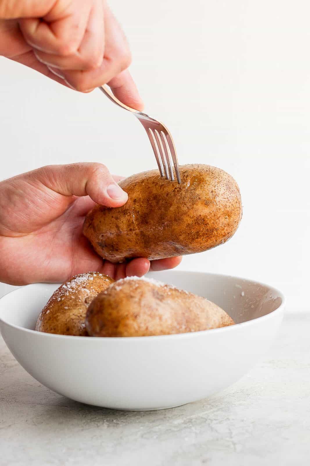 A hand holding a potato while another hand pierces it with a fork.