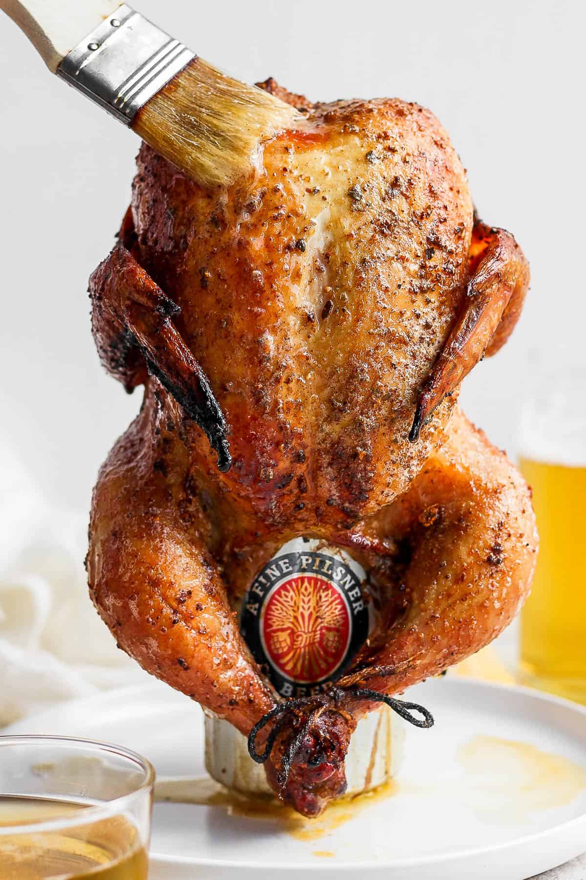 A fully cooked smoked beer can chicken on a plate as someone bastes with ghee. 