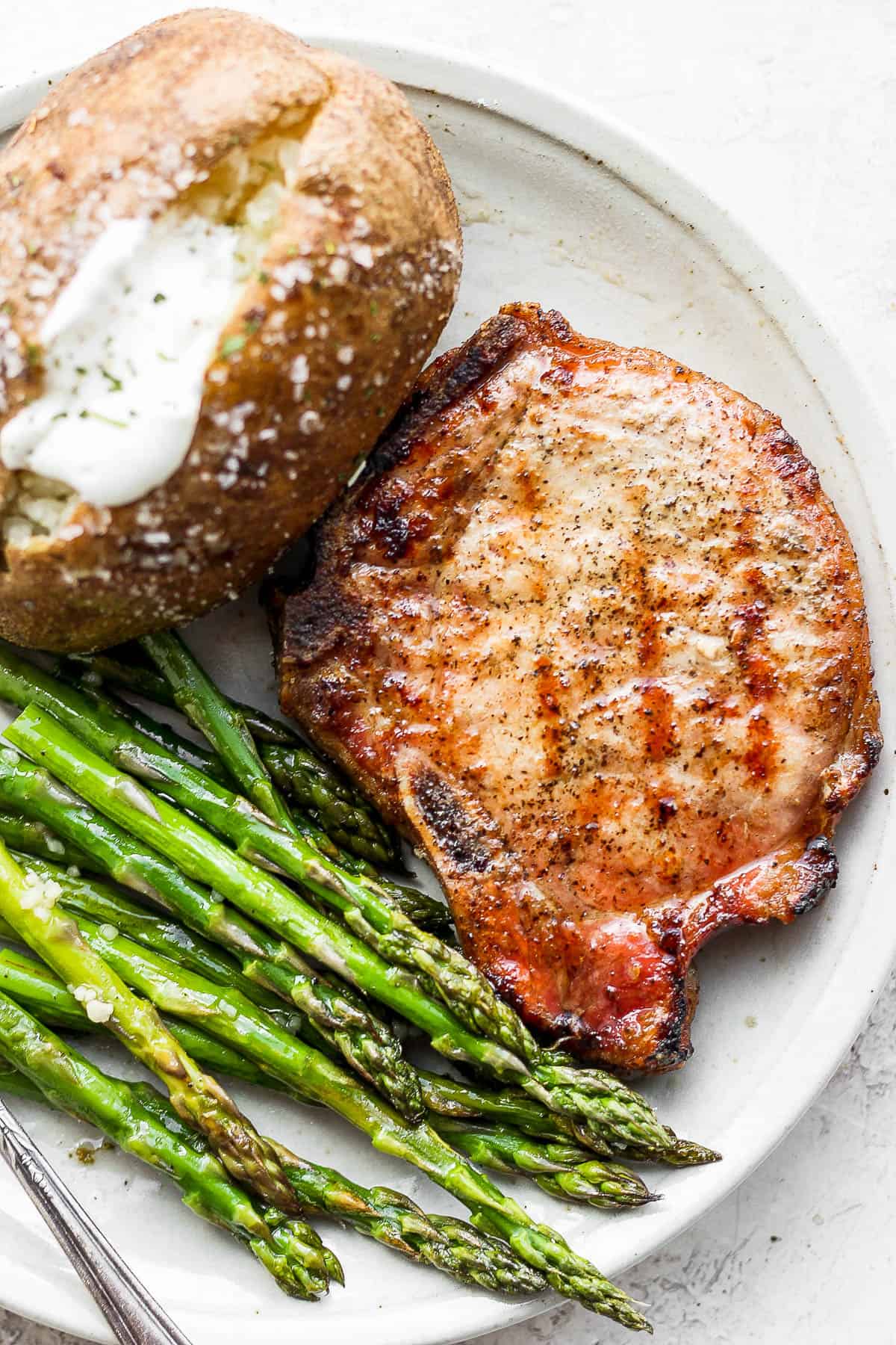 Plate with smoked pork chop, baked potato and asparagus on it. 
