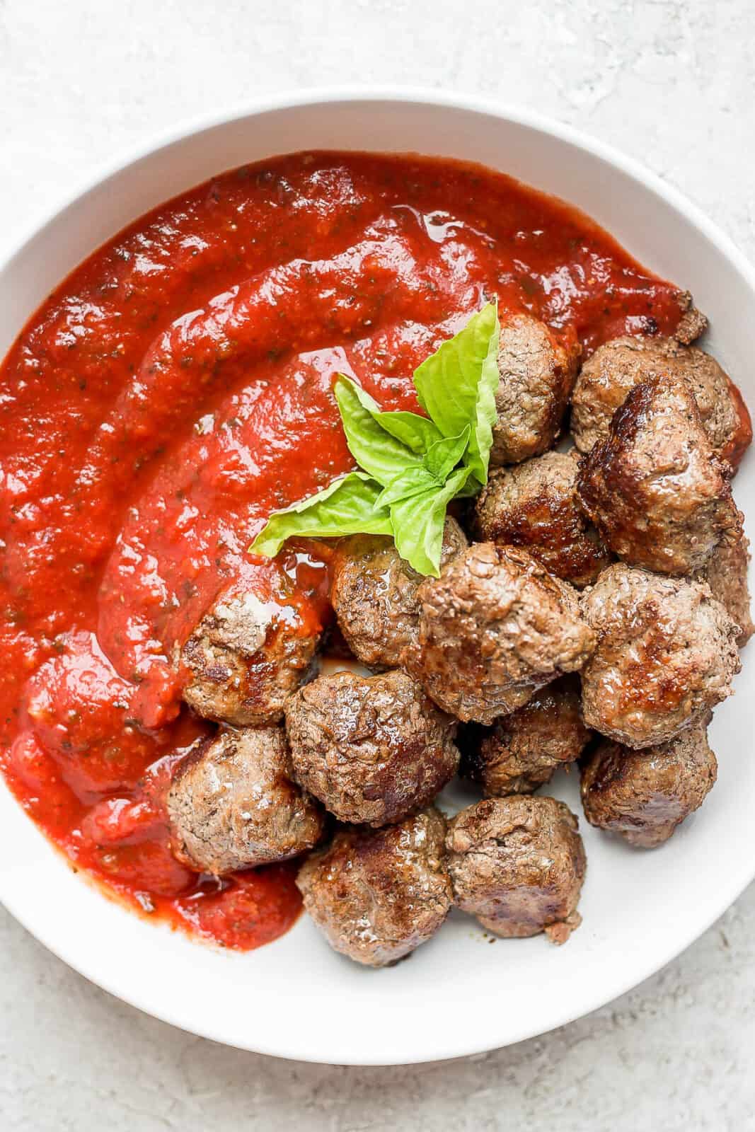 A bowl of cooked meatballs with pasta sauce and a little basil.