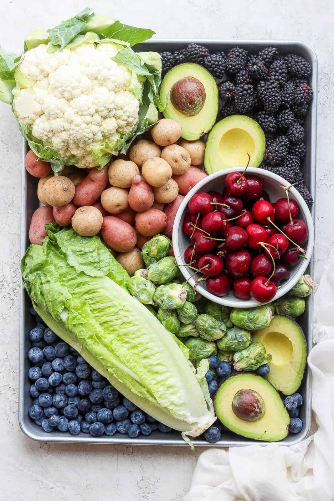 A baking sheet with cauliflower, blackberries, potatoes, avocado, cherries, brussel sprouts, romaine lettuce, and blueberries.