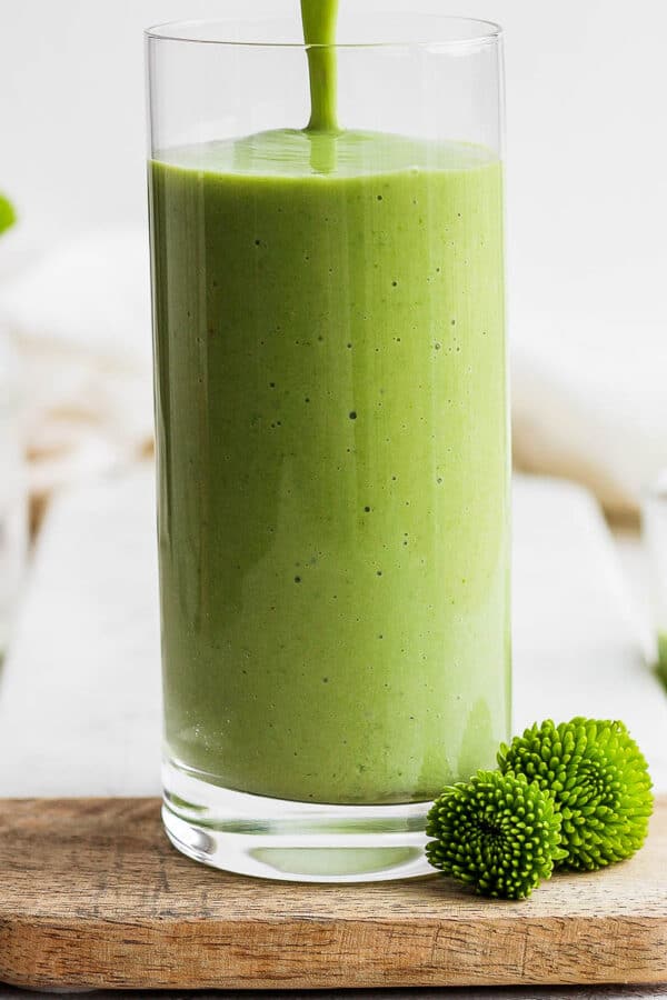 A green smoothie being poured into a glass with some green flowers.