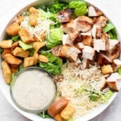 A large chicken caesar salad with a small bowl of caesar dressing.