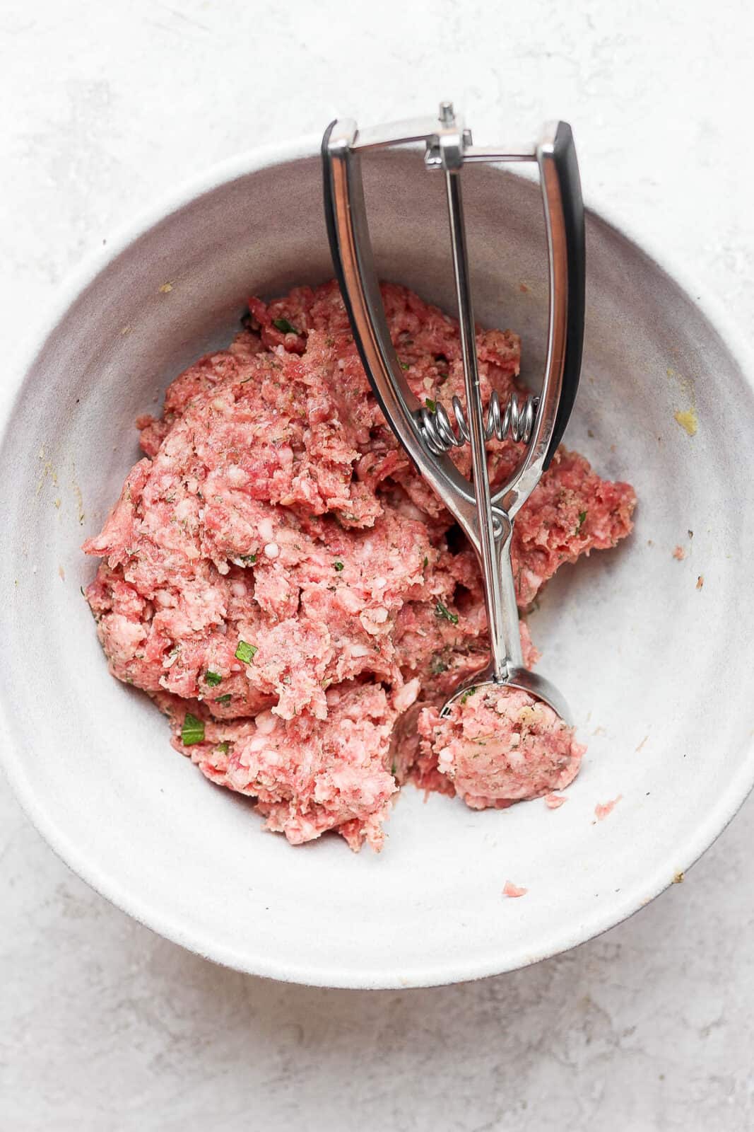 A bowl of lamb meatball ingredients all mixed together with a cookie dough scoop inside. 