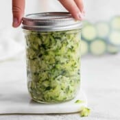 A jar of shredded zucchini with the lid being placed on top to go in the freezer.