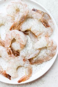 Raw, jumbo shrimp on a white plate seasoned with salt and pepper.