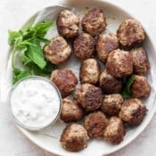 Several cooked lamb meatballs in a white dish with some tzatziki sauce in a bowl and fresh mint.
