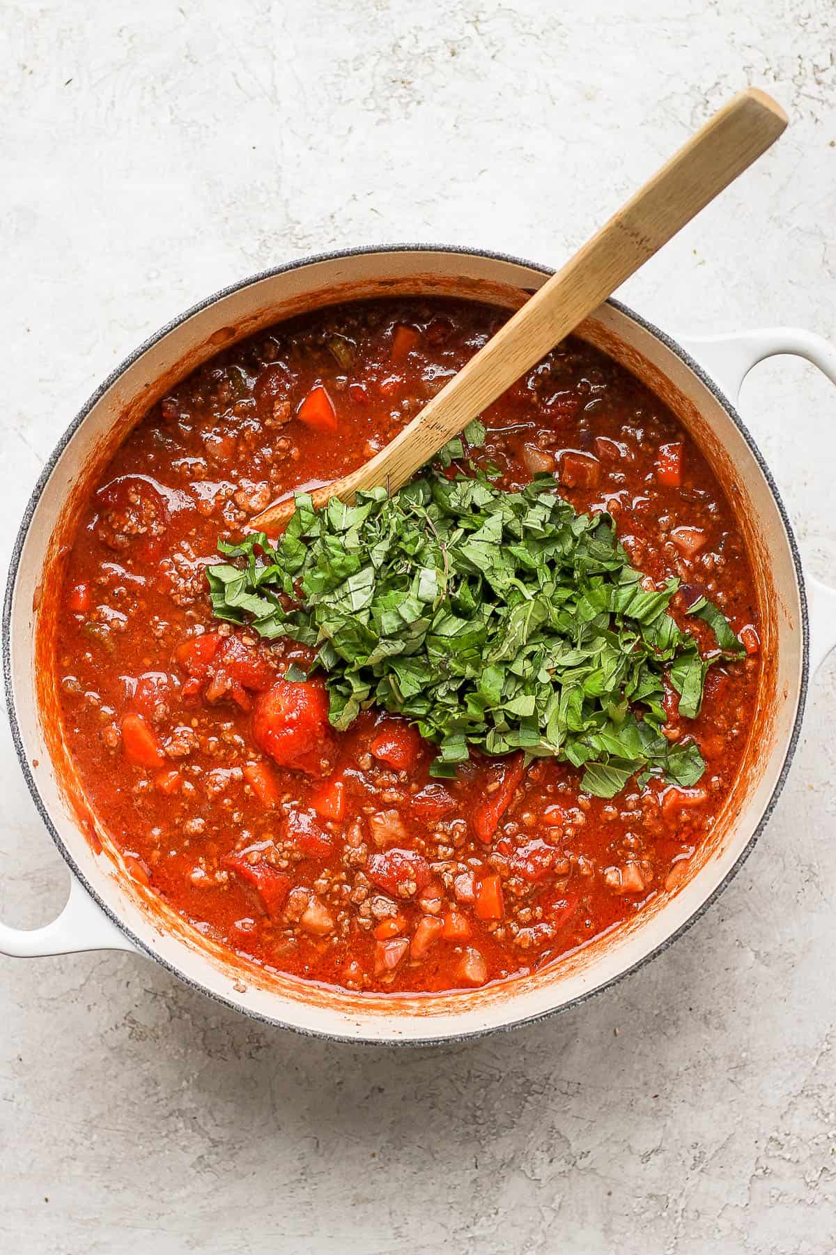 Dutch oven full of bolognese with fresh herbs on top and a wooden spoon sticking out.