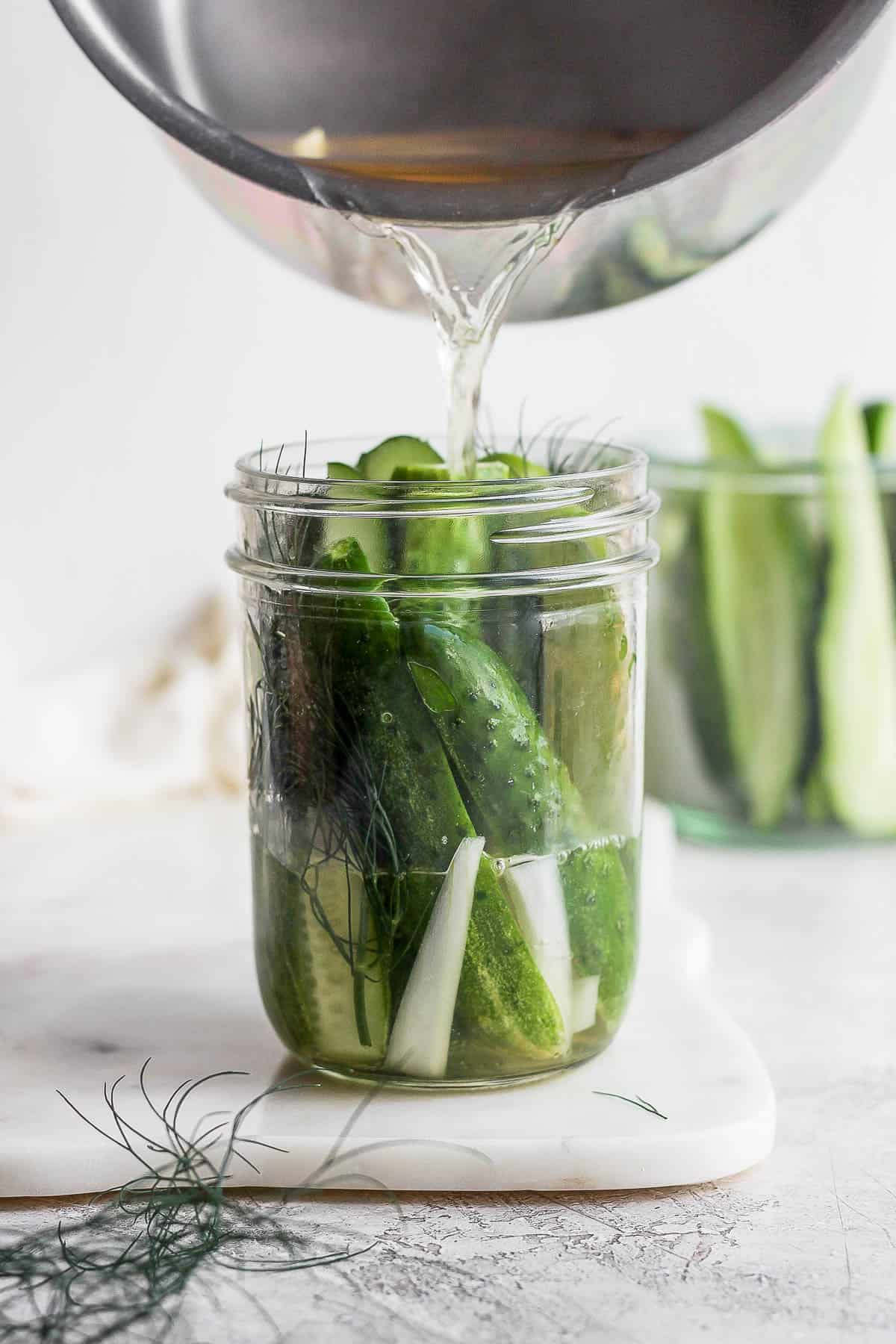Pickling brine being poured into a jar with sliced cucumbers, dill, and onion to make easy refrigerator dill pickles.