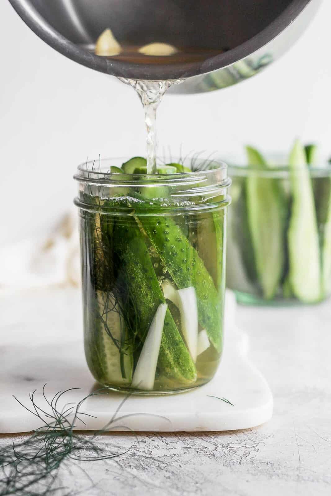 Someone pouring hot brine into a mason jar full of cucumbers, onions and dill. 