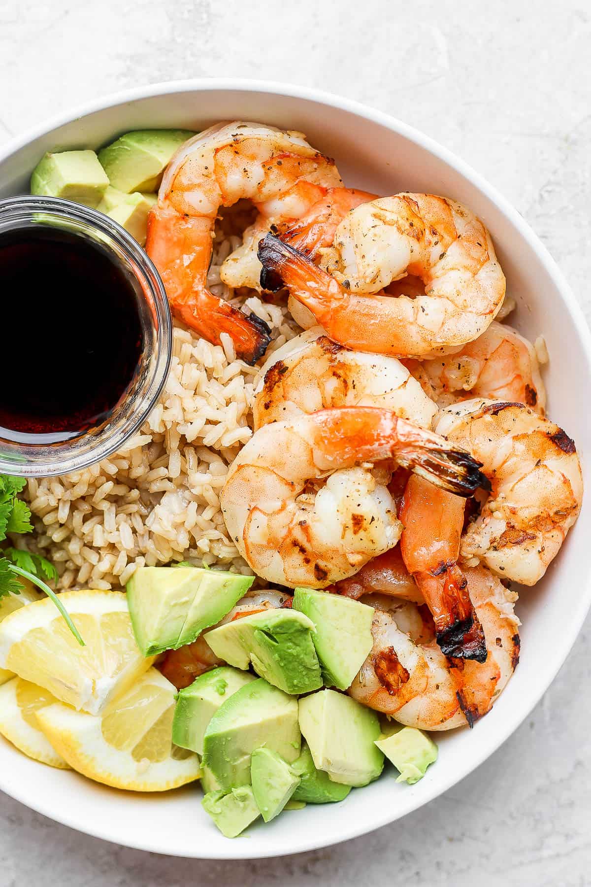 Pan fried shrimp in a bowl with rice, avocado, and a dish of soy sauce.