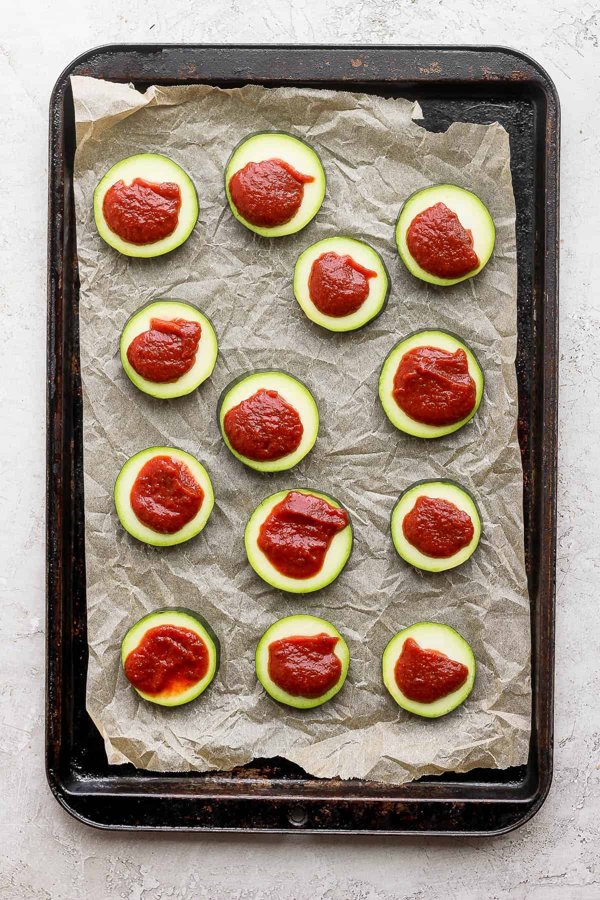 Sliced of zucchini with pizza sauce on top on a parchment lined baking sheet.