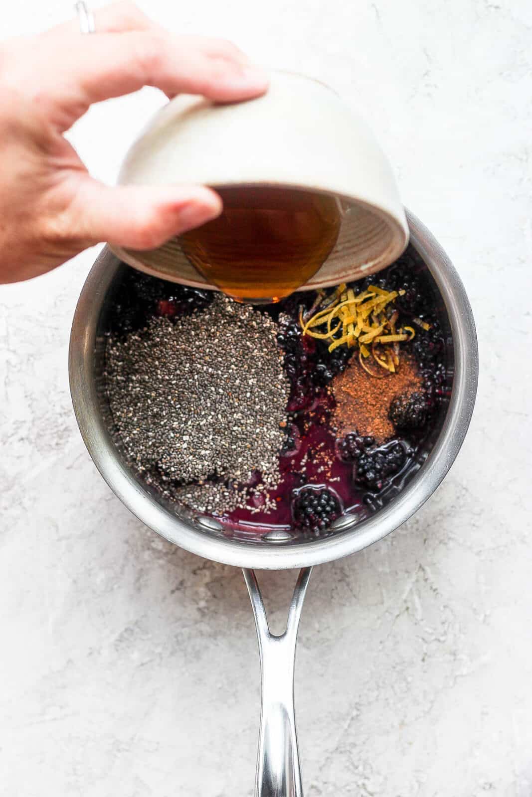 Someone pouring maple syrup into a saucepan filed with other chia jam ingredients.